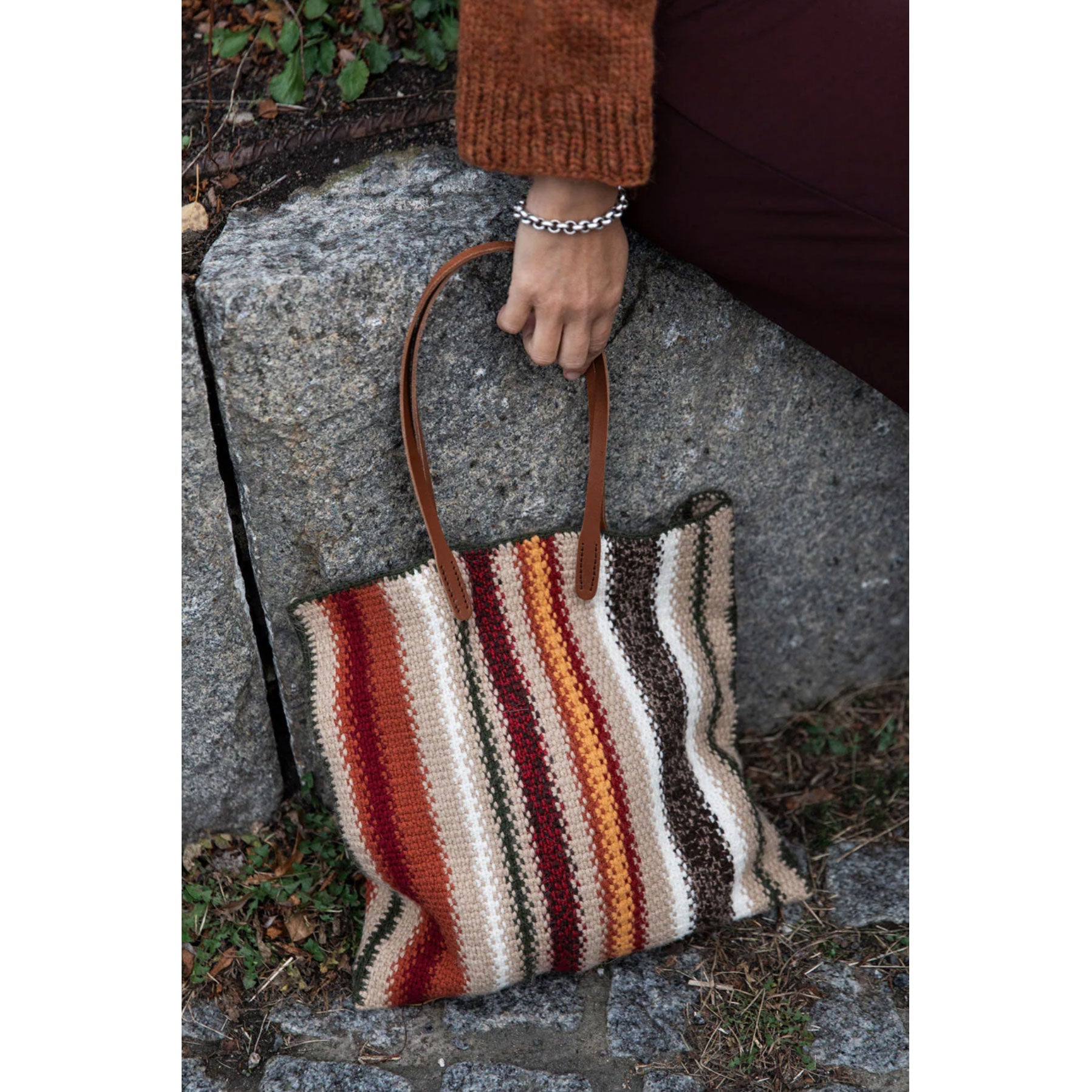 Striped handbag with brown handle held by a person on a stone surface