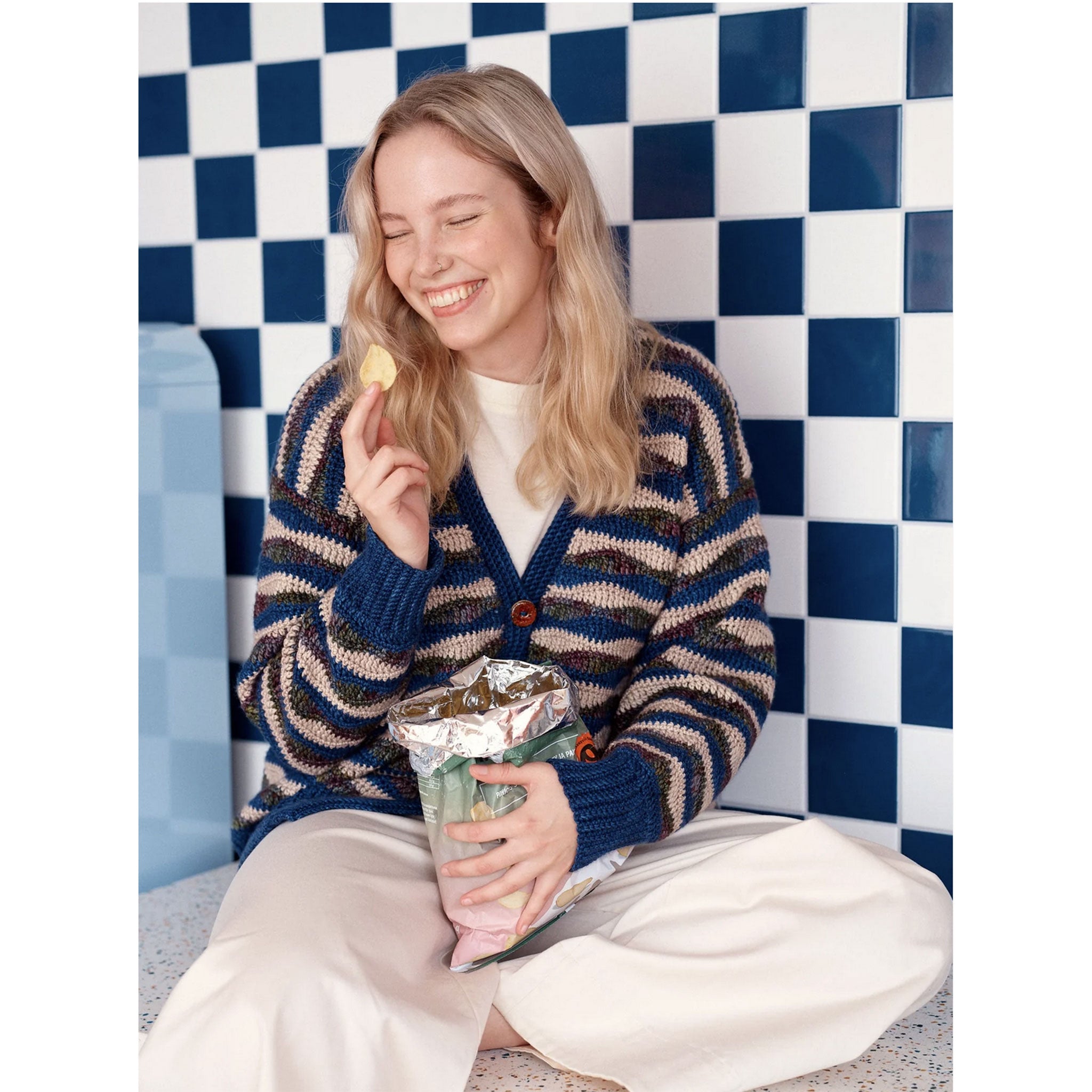 Woman wearing a striped crocheted cardigan sitting on a checkered floor holding a bag of chips and a container.