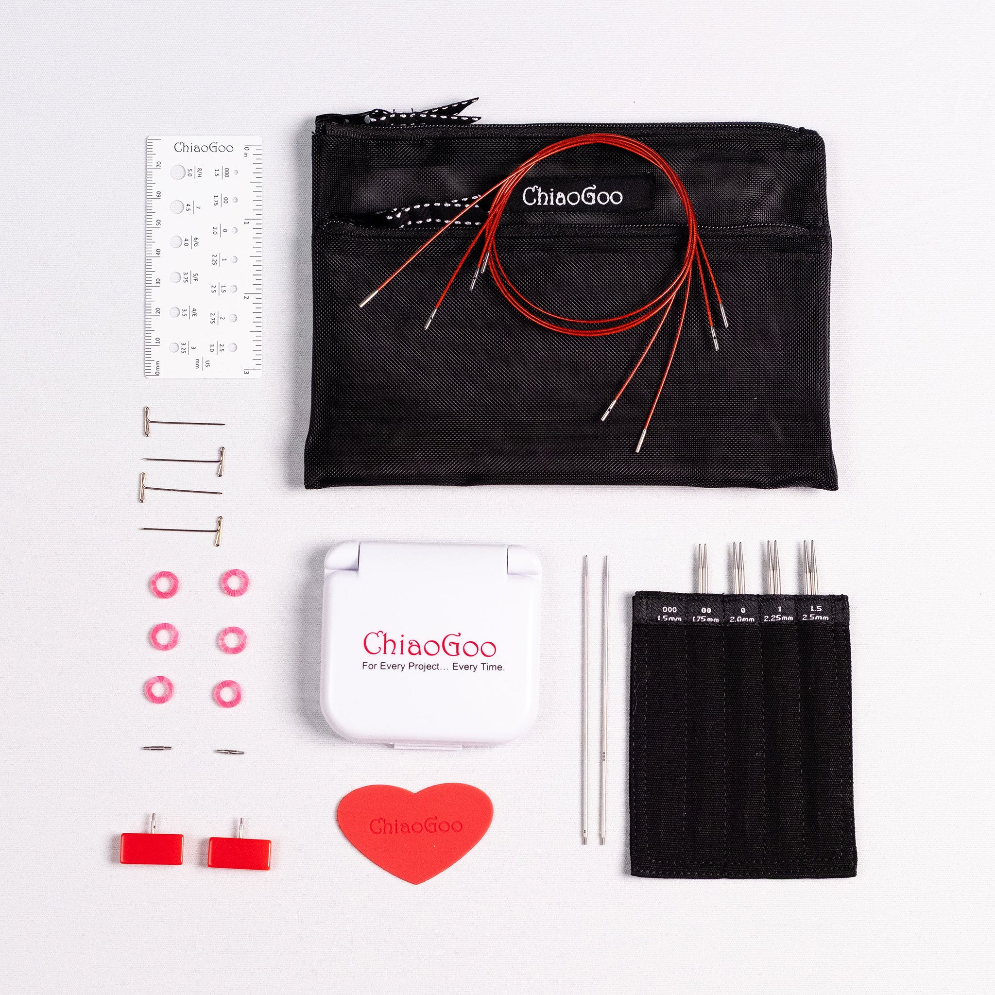 ChiaoGoo 4in Mini set showing labelled tip sleeve with screw ends of tips showing, three red lace cables lying coiled, a white plastic ruler needle gauge and white plastic click shut container with red ChiaoGoo logo, red rubber heart, two red mini end stoppers, six pink stitch markers, two MINI T-pins and two MINI cable connectors