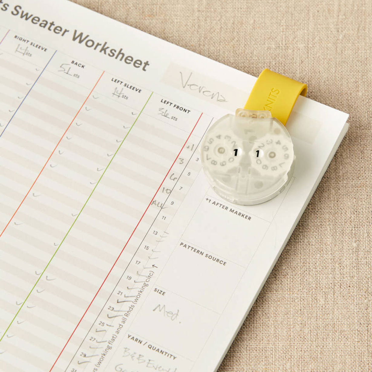 Knitting row counter magentically attached to yellow makers clip on a sweater worksheet with measurements
