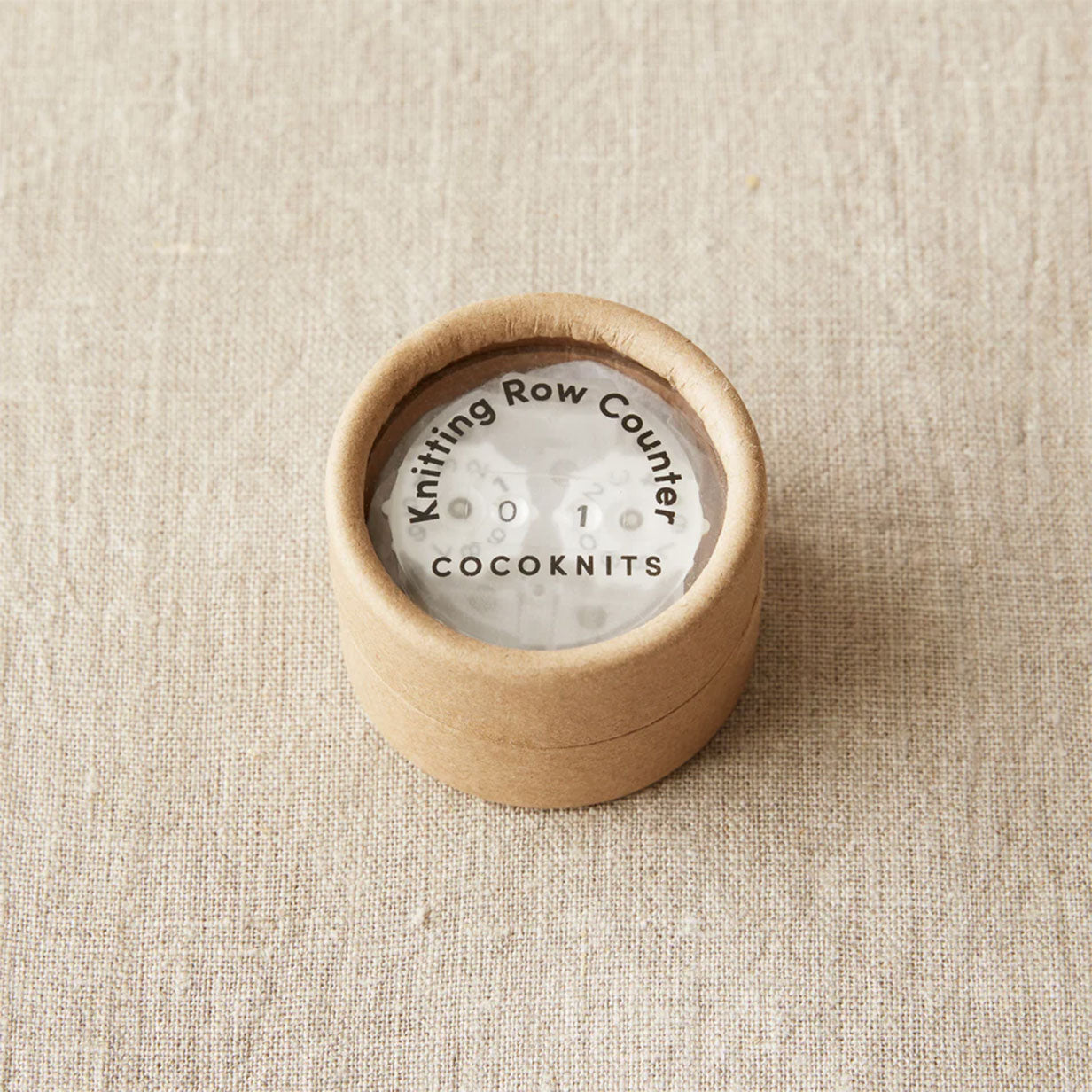Knitting row counter in a cardboard kraft coloured pot on a linen fabric background