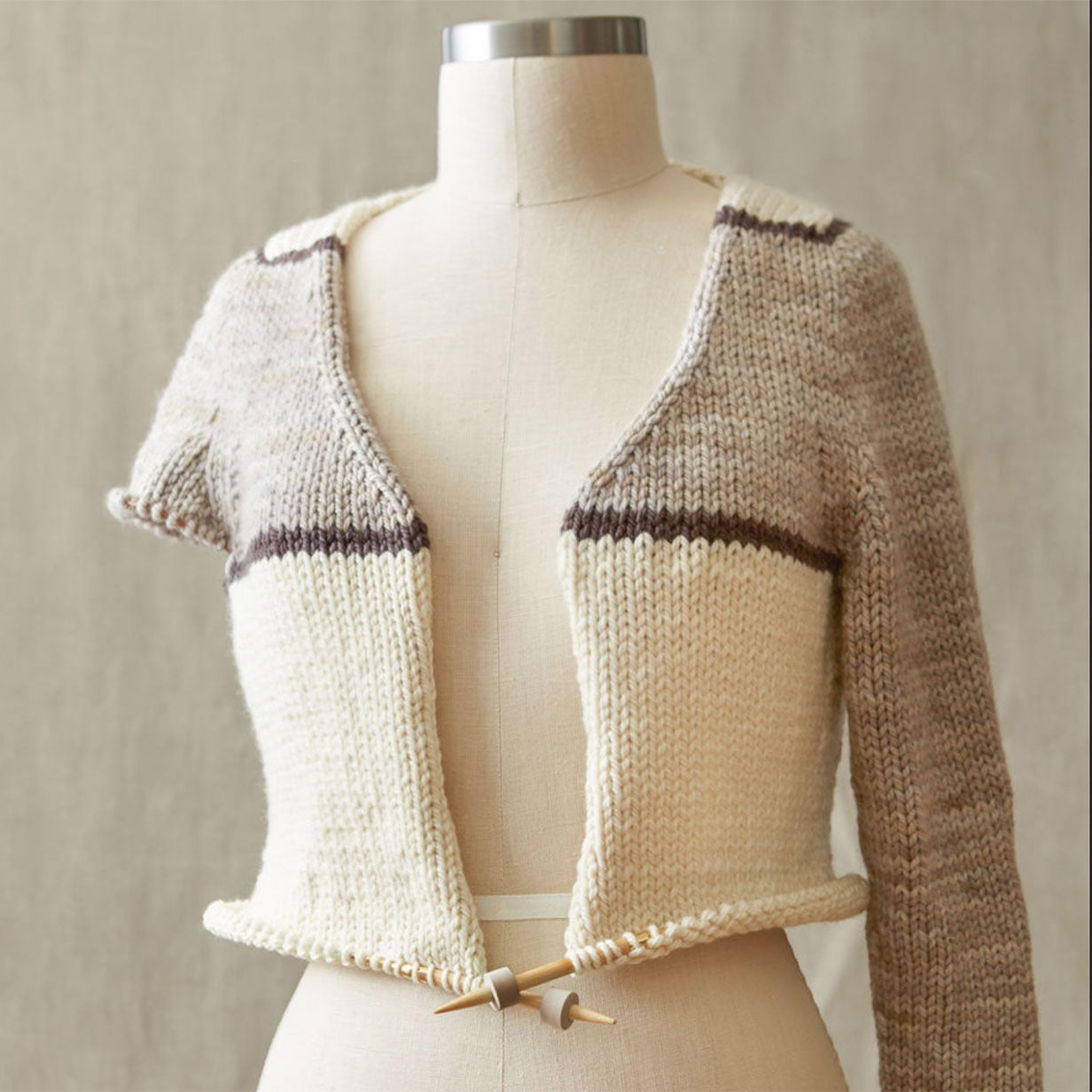 Knitted cardigan on a mannequin against a neutral background, stitch stoppers placed on the end of the wooden needle tips