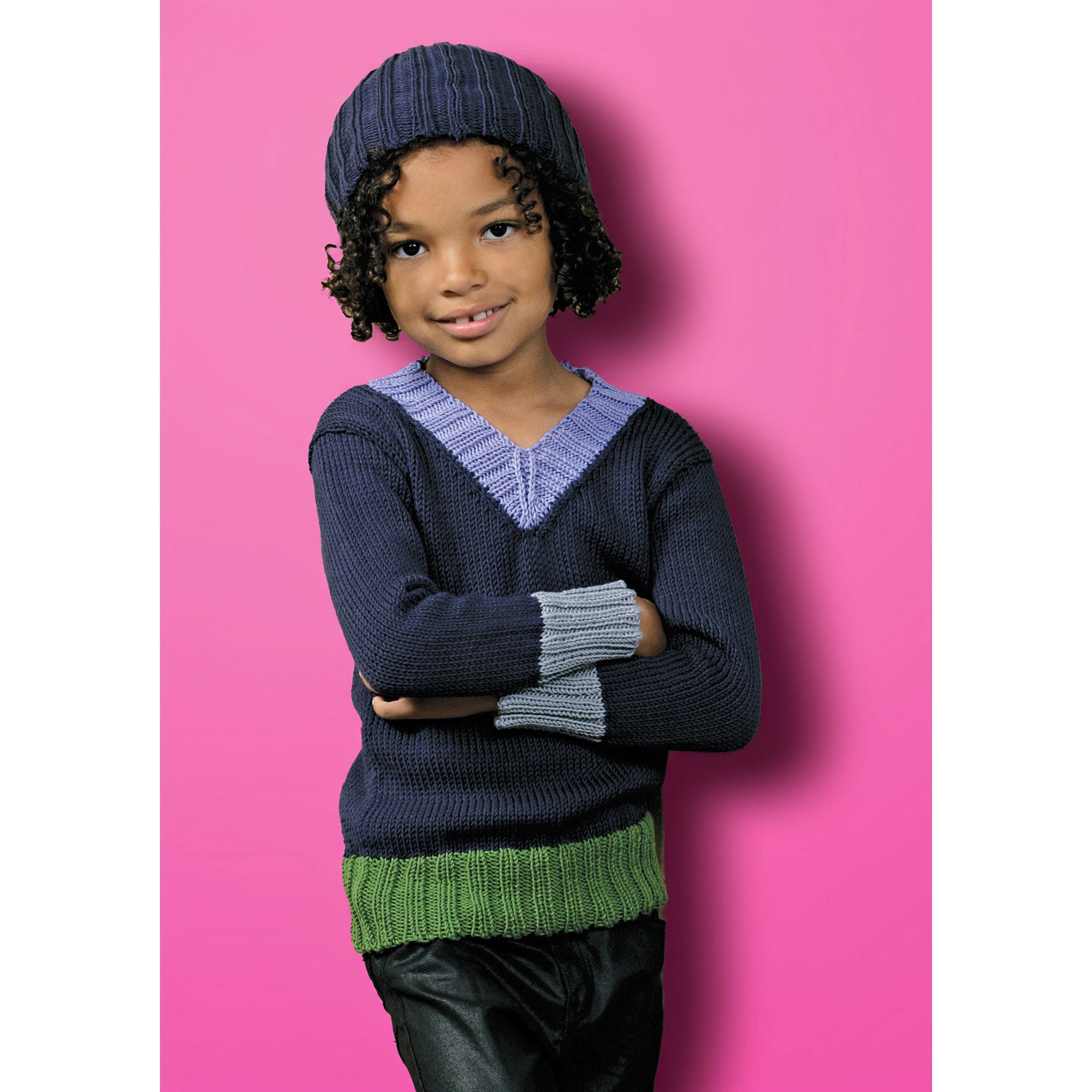 Child wearing a navy sweater with green trim and a blue knit hat against a shocking pink background