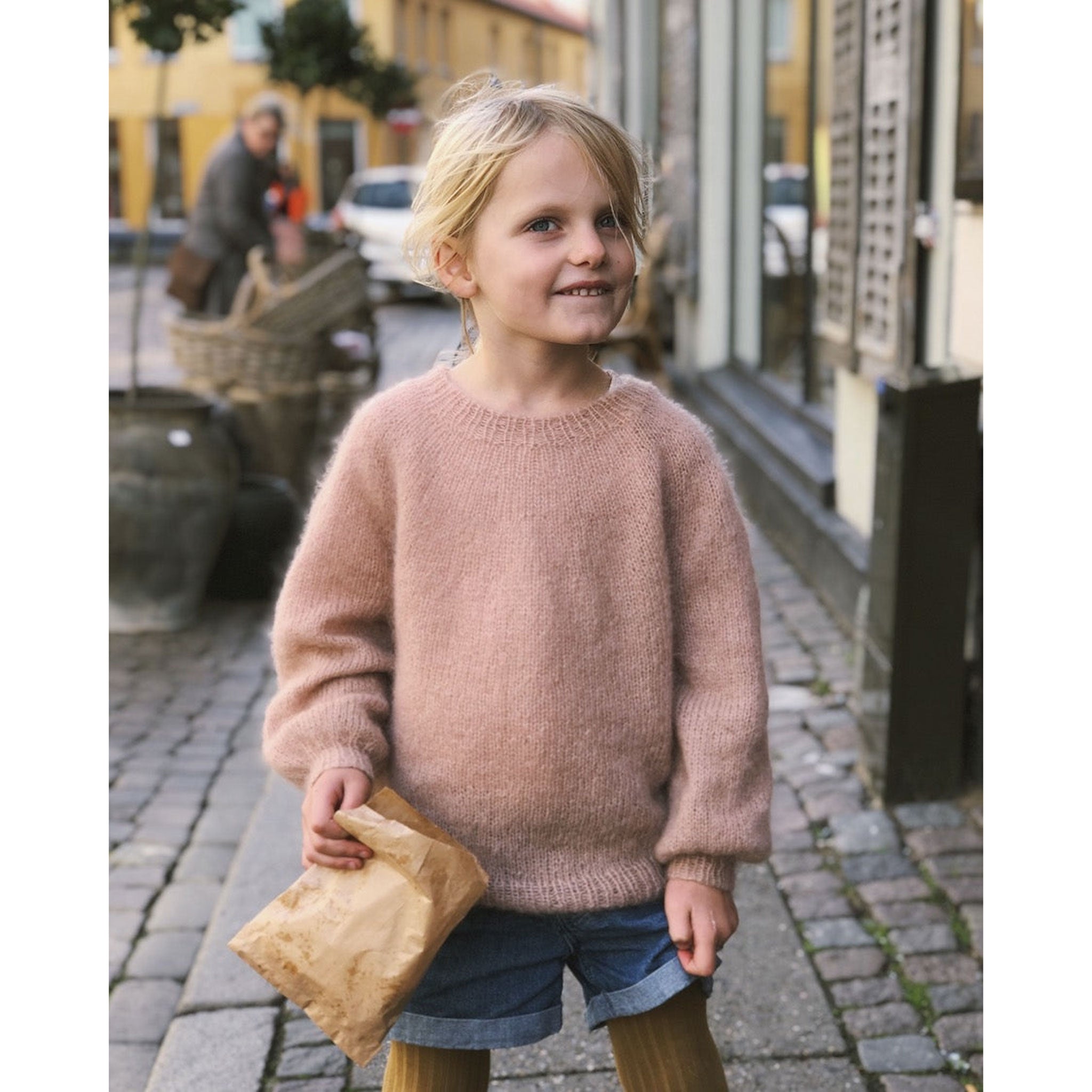 Young girl in a pink knitted sweater standing on a street holding a brown paper bag.