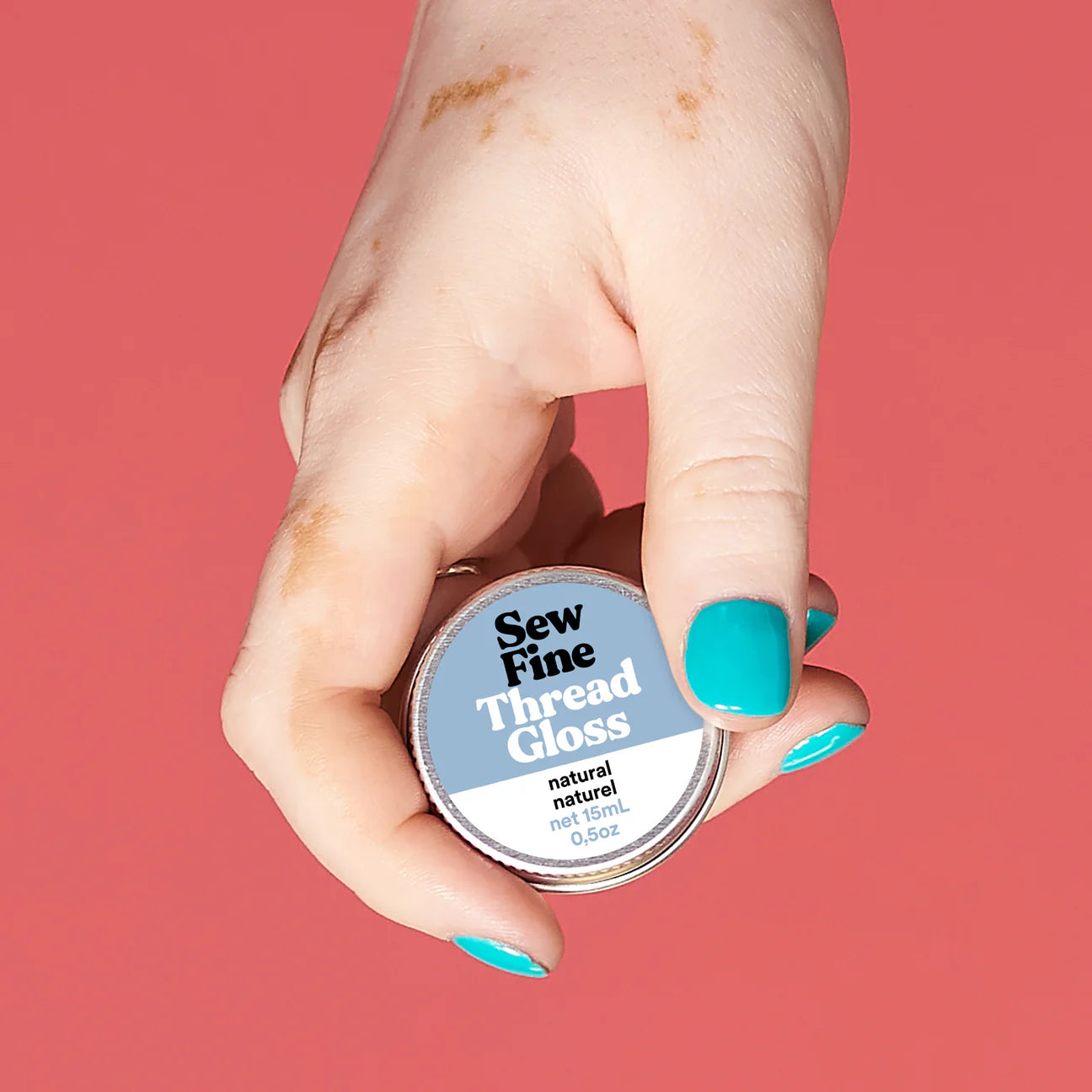 Hand holding a labelled tin of Sew Fine Thread Gloss - Natural against a coral colour background