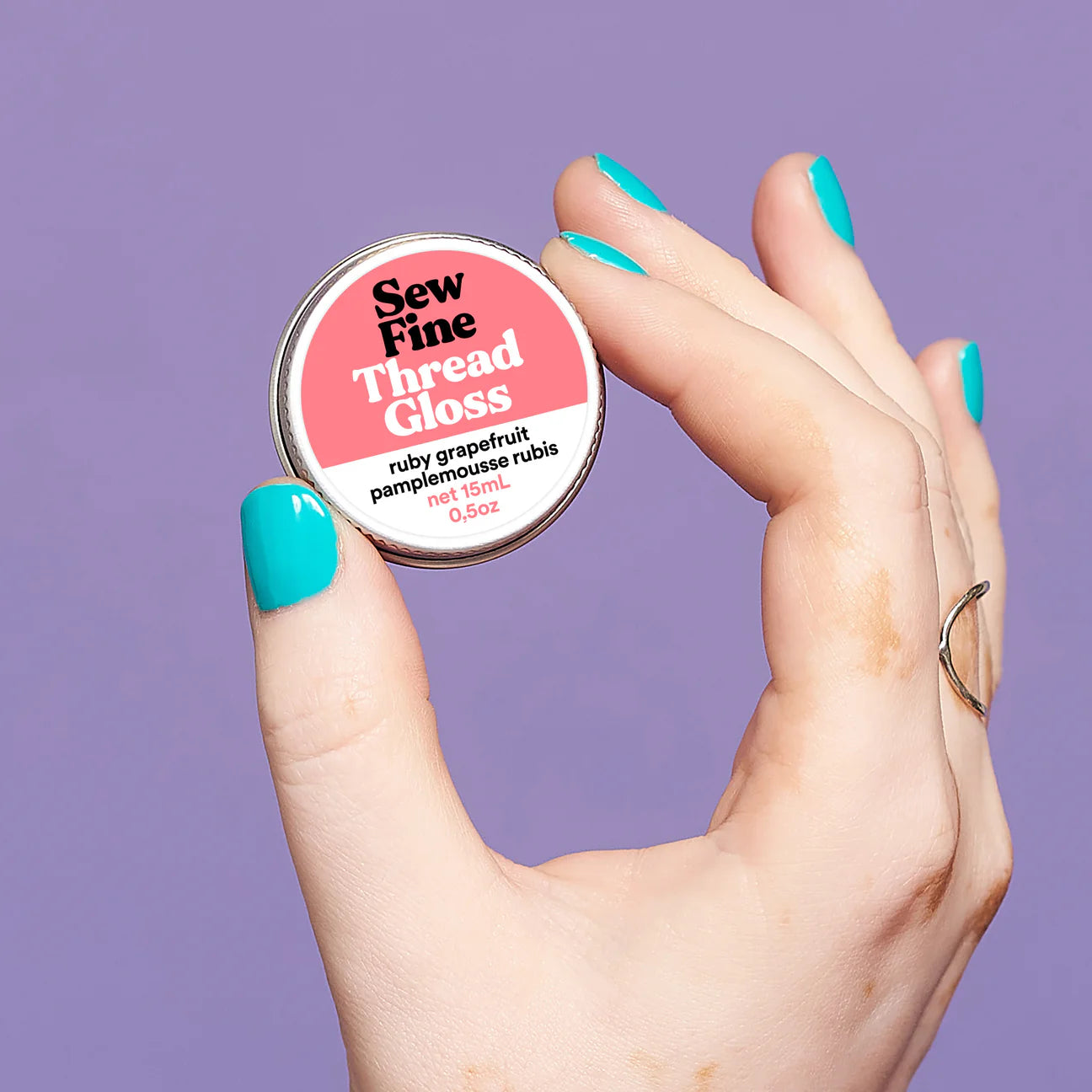 Hand holding a labelled tin of Sew Fine Thread Gloss - Ruby Grapefruit against a lilac background