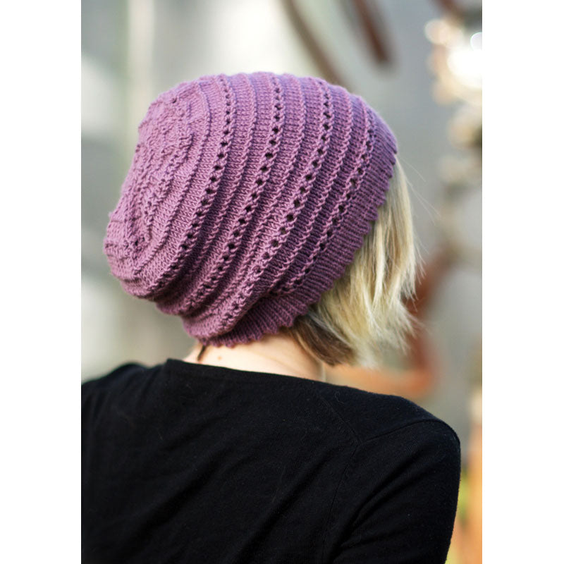 Back view of person wearing purple coloured knitted slouch hat with picot rim, garter stitch contrast and eyelets, against a blurred background