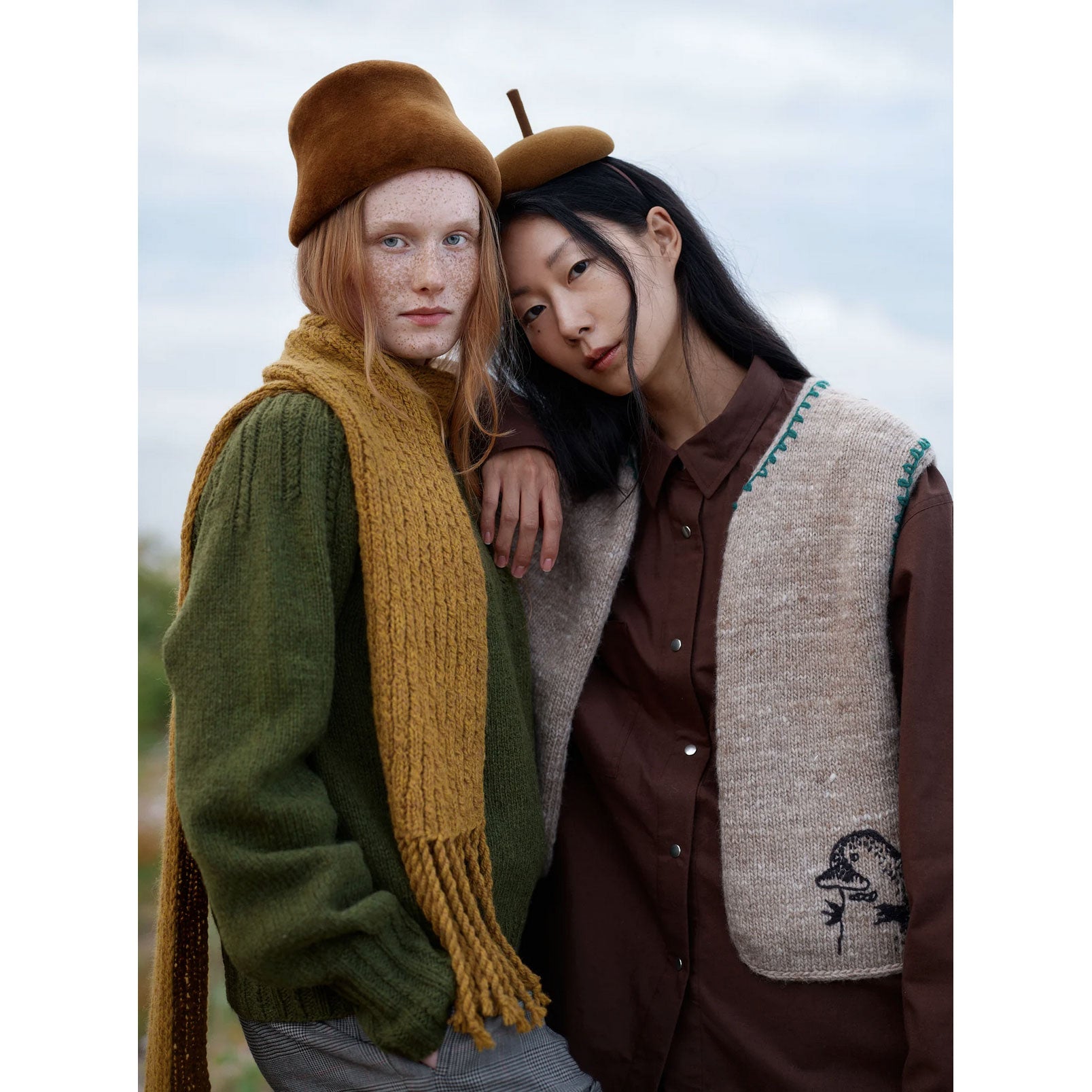 Two people wearing autumn-themed clothing with scarves and hats against a blurred natural background.