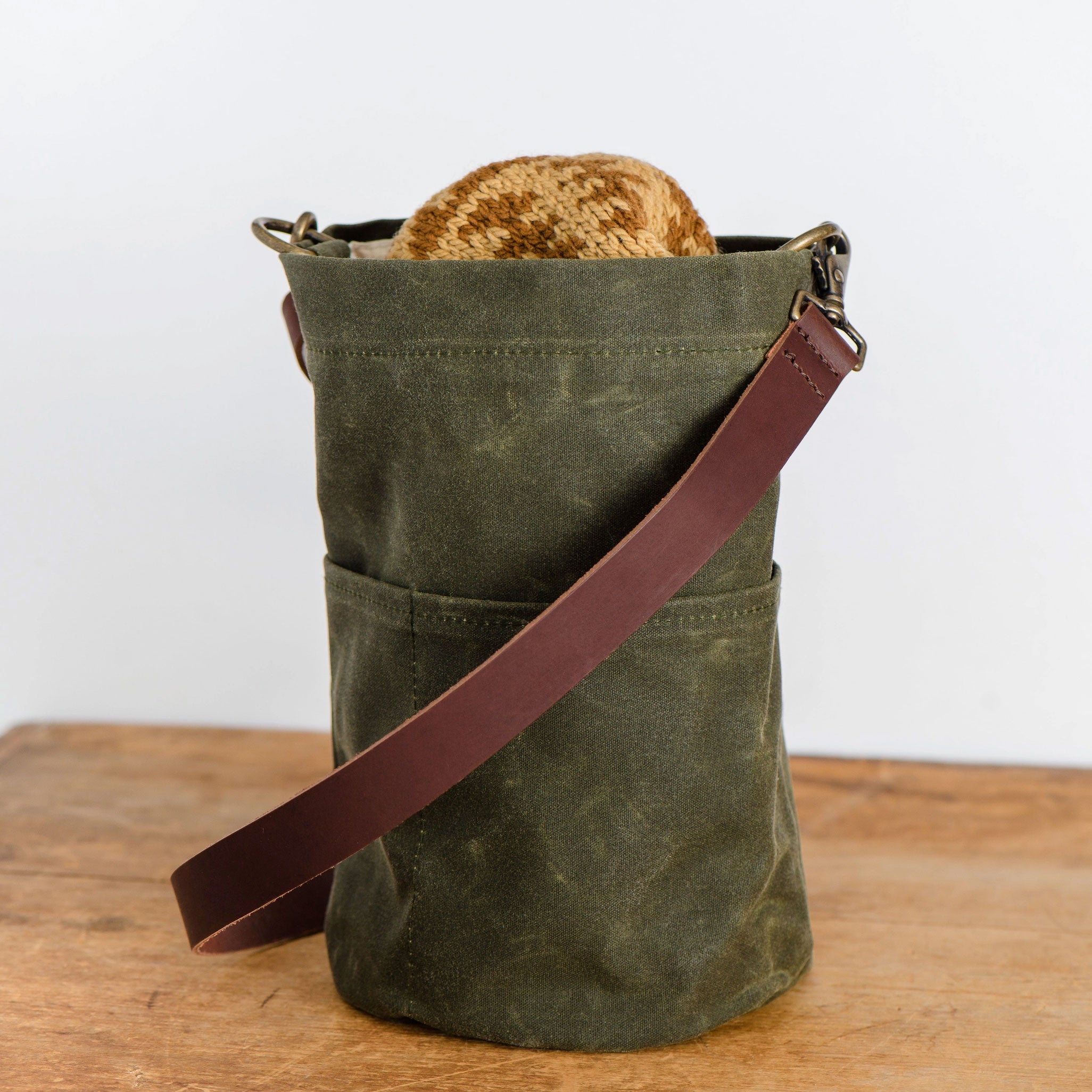 Olive green canvas bag with brown leather strap with brown knitting project showing on a wooden surface.