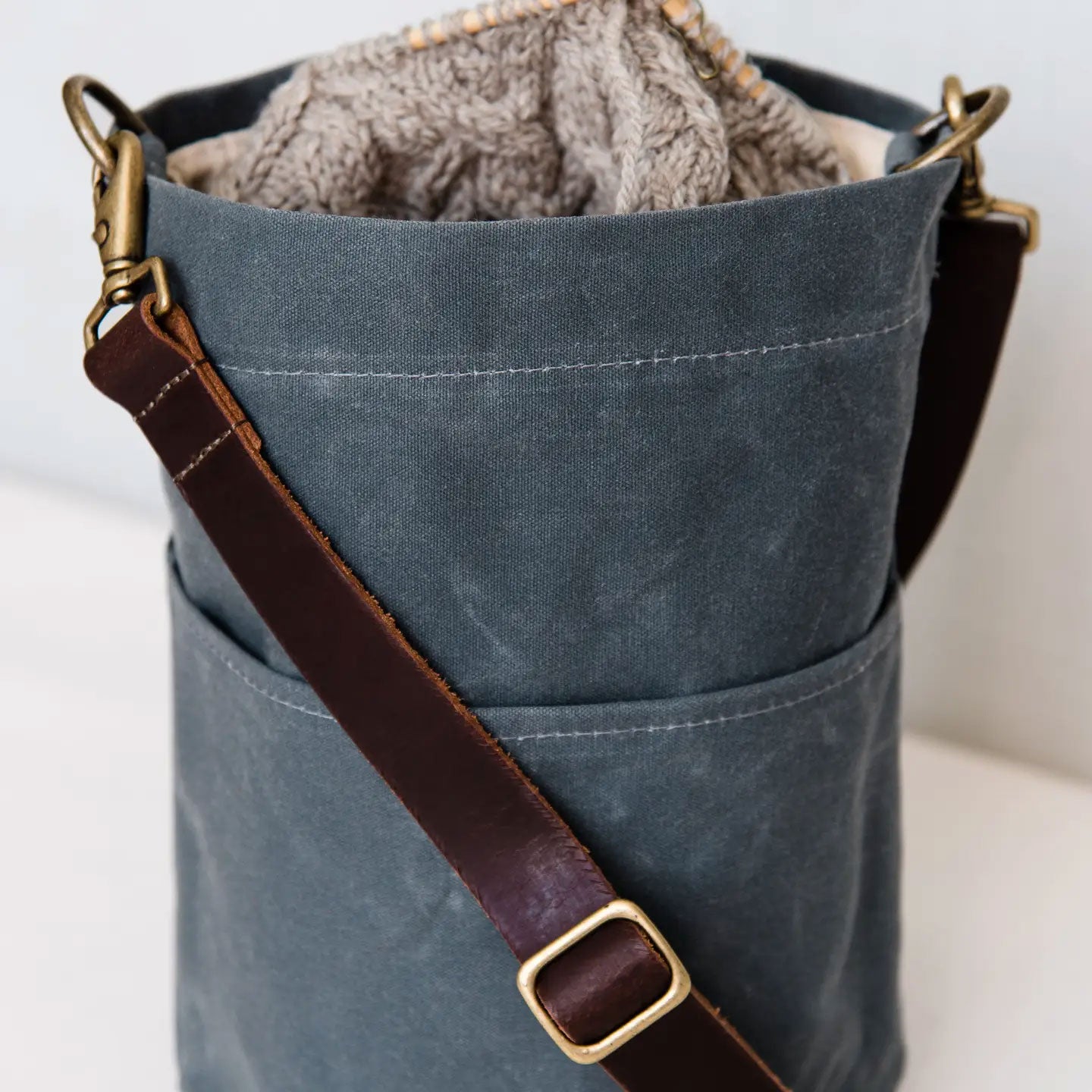 Slate blue canvas bag with brown leather strap, grey knitting project showing and brass buckle on a white background