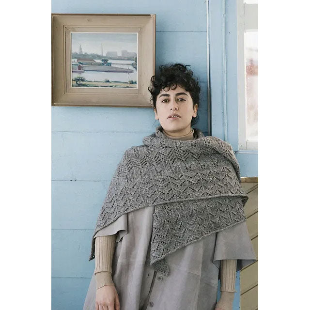Aloft by Bekah Knits. Woman wearing a grey knitted triangular shawl against a light blue wall with a framed picture.
