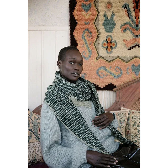Crosshatch by Rachel Brockman. Person wearing a large green and cream knitted triangular patterned scarf in a room with a decorative rug.