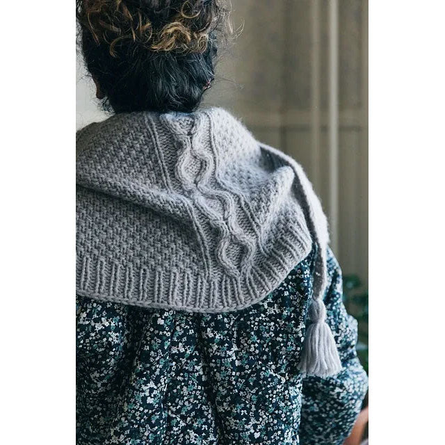 Sulani by Stephanie Earp. Person wearing a gray knitted shawl with double moss stitch and cables, standing indoors.