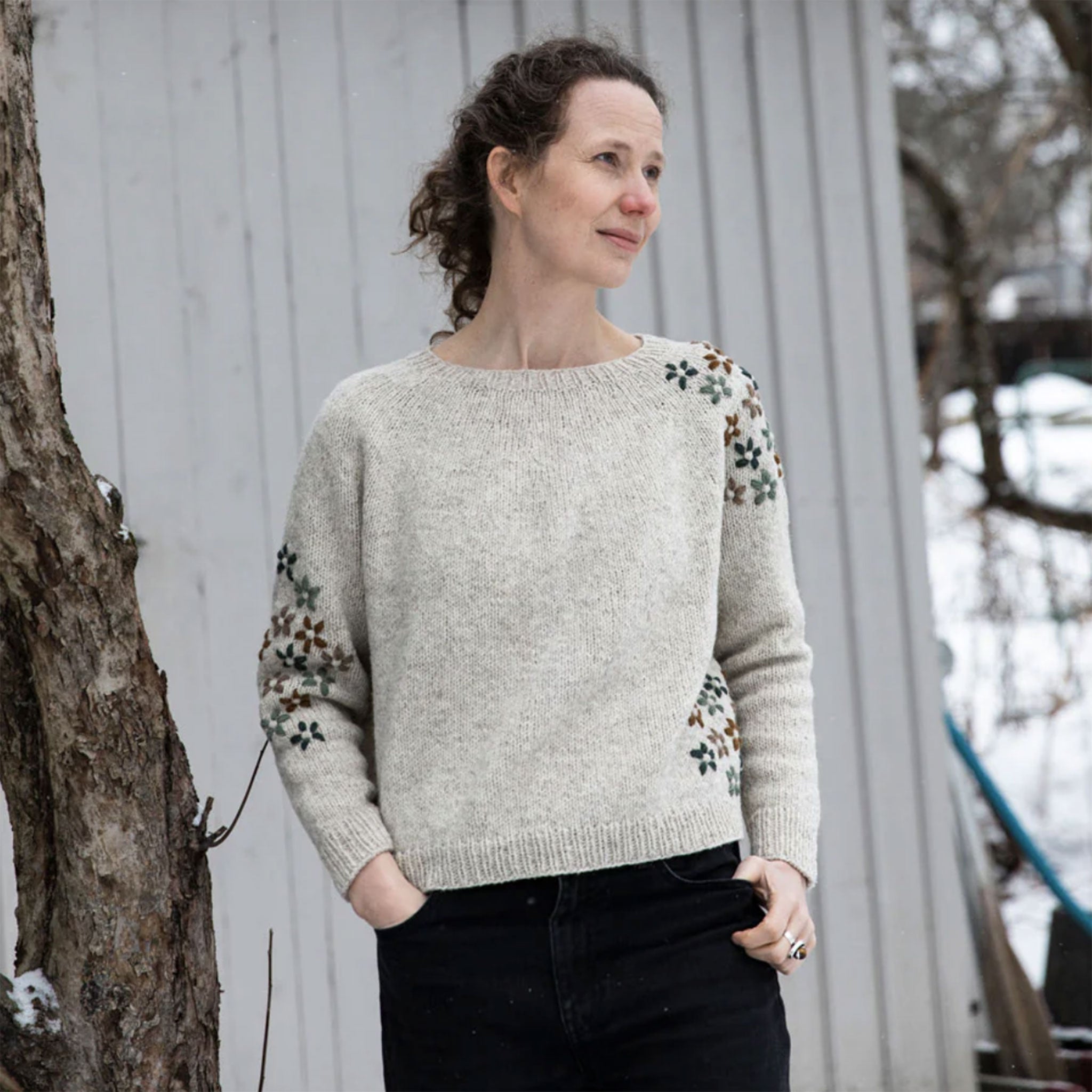 Woman wearing a hand knitted light gray sweater with embroidered flowers on sleeves outside with shed wall behind
