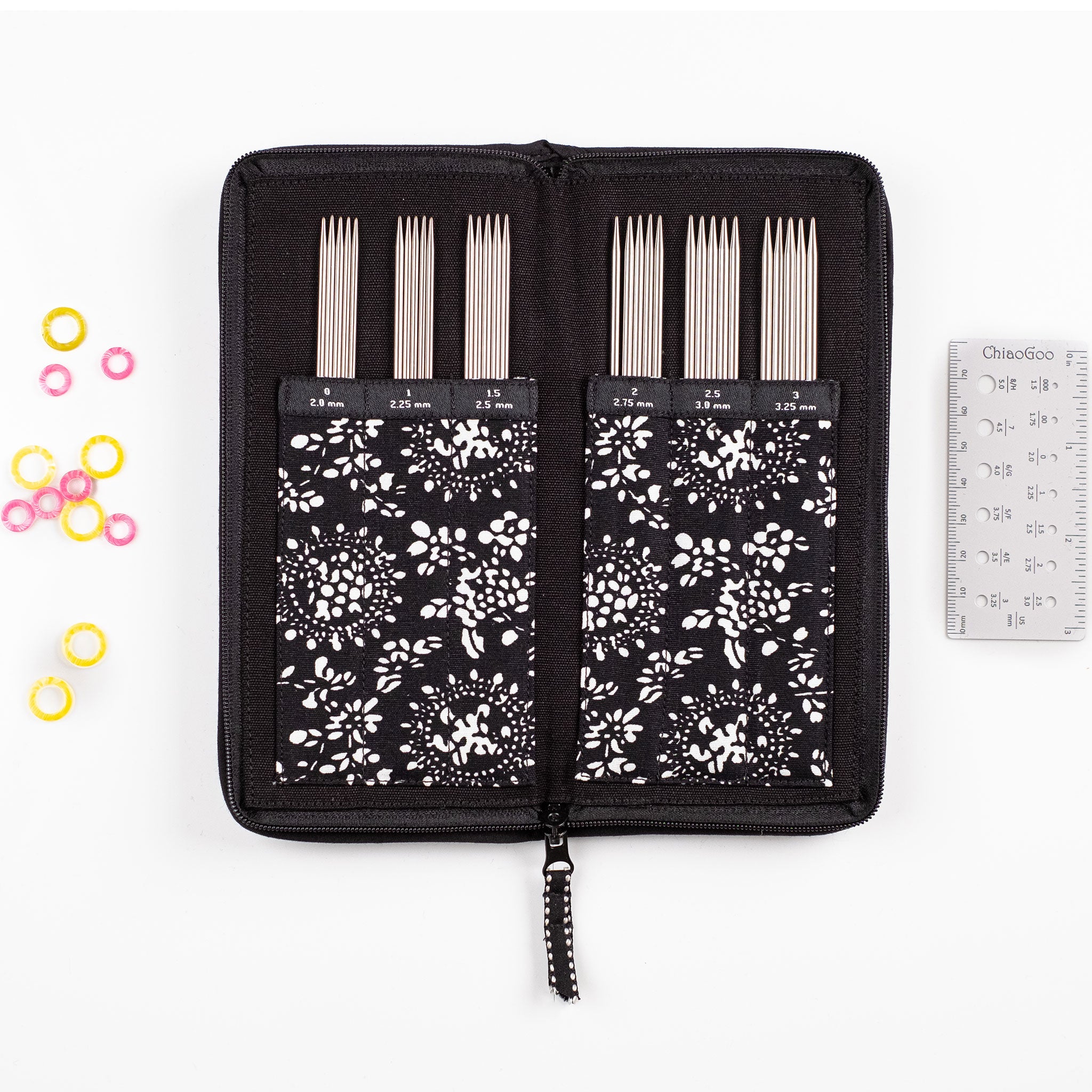 Set of stainless steel DPNs in a black fabric case with floral pattern, surrounded by colourful stitch markers and a white needle gauge ruler on a white background.