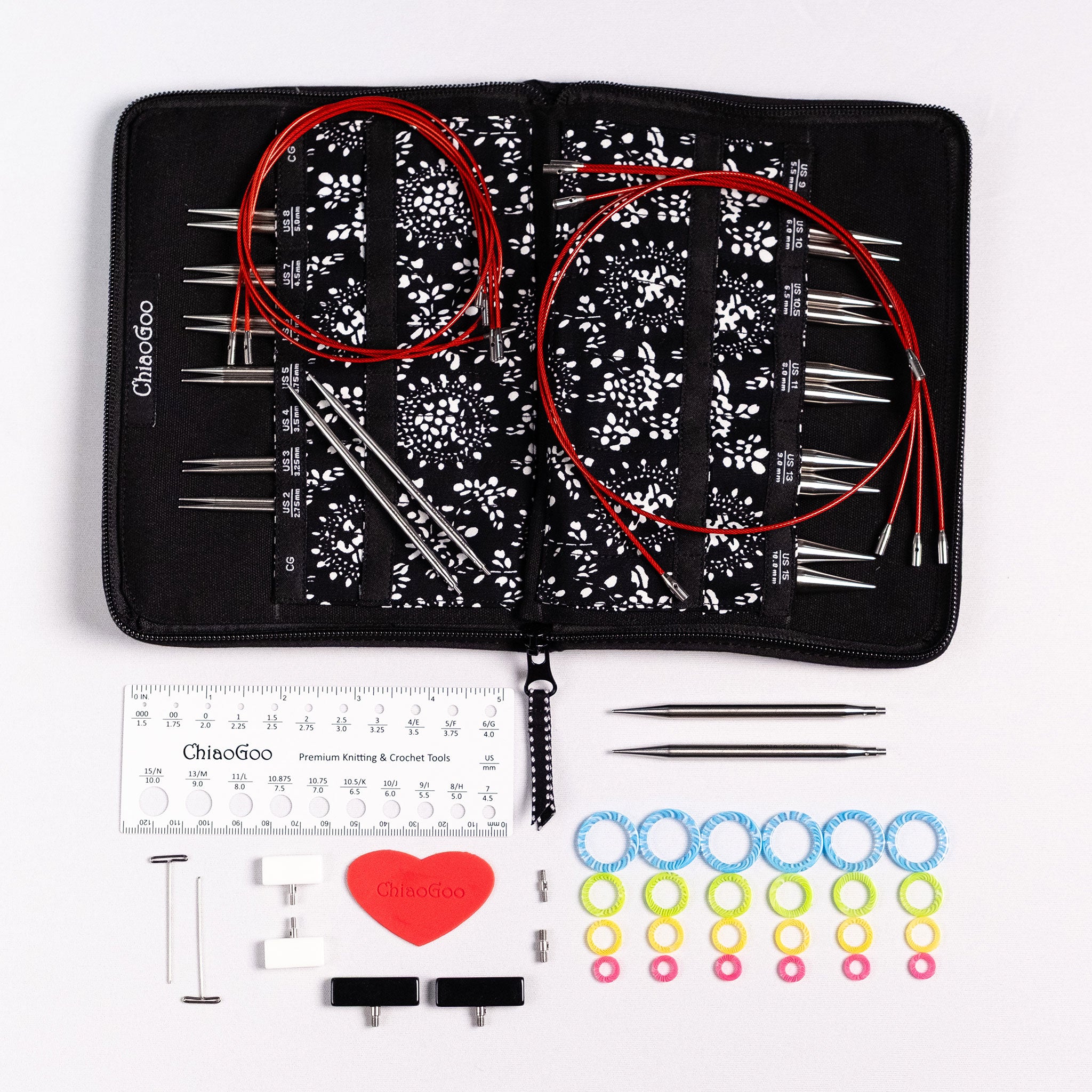 Complete set of ChiaoGoo 4in long stainless steel interchangeable knitting needles showing black zippered case lying flat and open with tips inserted in labelled pockets, red lace cables coiled on top, the included accessories displayed in front of the open case, on a white surface