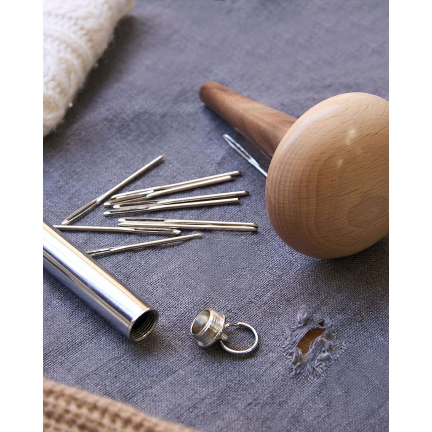 Set of darning needles next to silver coloured storage cylinder and wooden darning mushroom on a blue woven textured surface