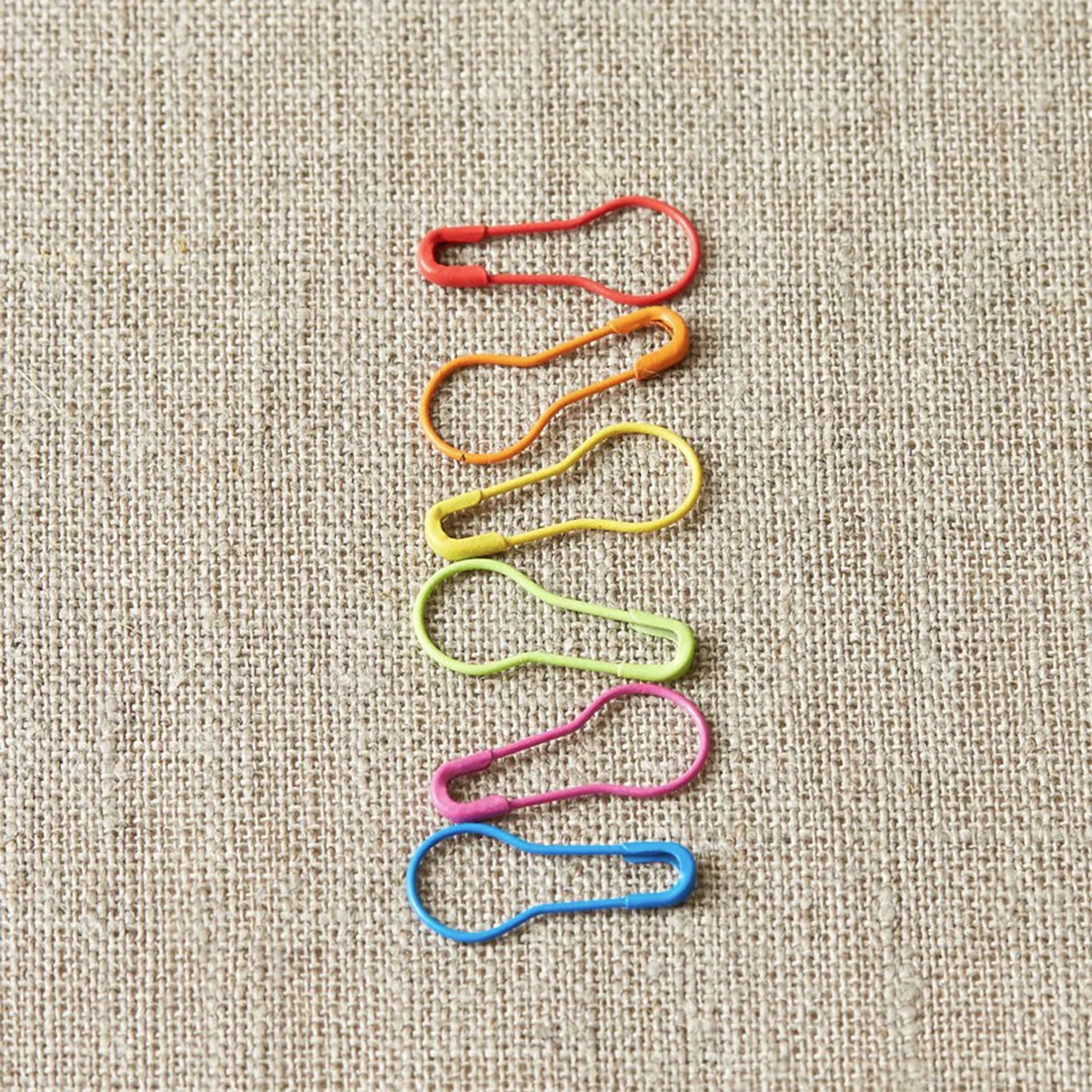 Six Cocoknits colourful opening bulb style stitch markers, six, in a column, red, orange, yellow, green, pink and blue on a linen background