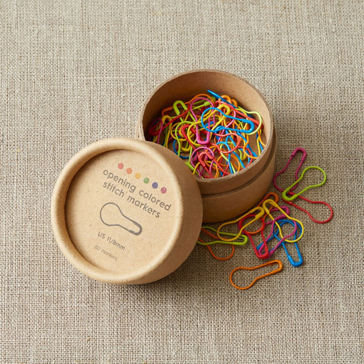 Open kraft coloured cardboard storage pot with colourful bulb style stitch markers on a textured linen surface