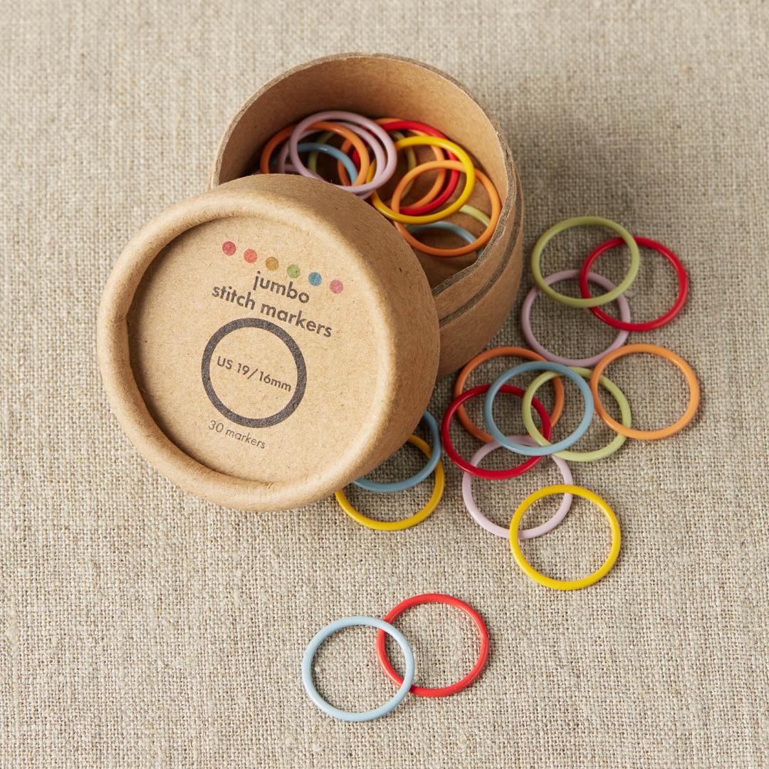 A small cardboard storage tub with jumbo fixed colourful stitch markers spilling out and onto linen cloth in pale blue, red, yellow, pale green, light orange and pale pink
