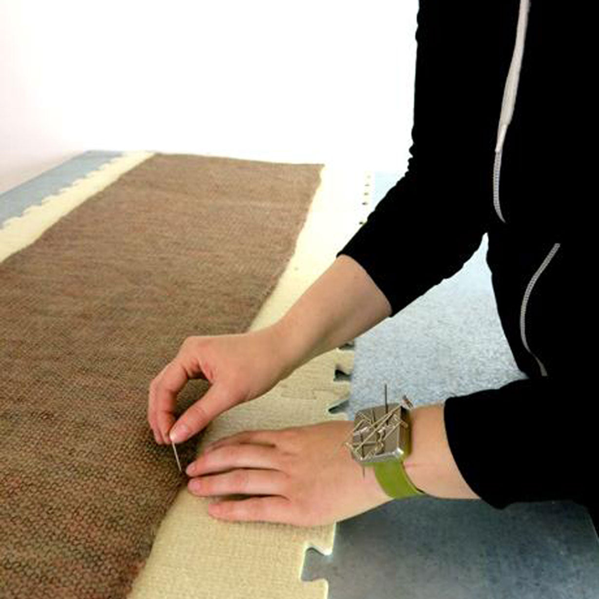 Close-up of person pinning a long knitted scarf into fibre covered EVA blocking mat with a T-pin, the person is wearing a green wrist band with a magnetic face and a pile of T-pins stuck to it