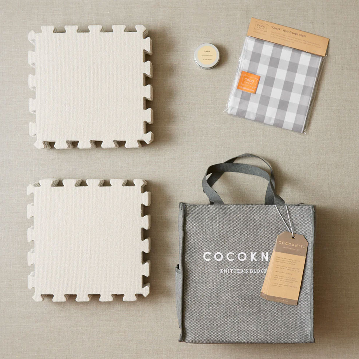 A flat lay of two piles of EVA interlocking mats, a tin of T-pins, a gingham measuring cloth and a jute storage bag showing the Cocoknits logo on a plain linen background