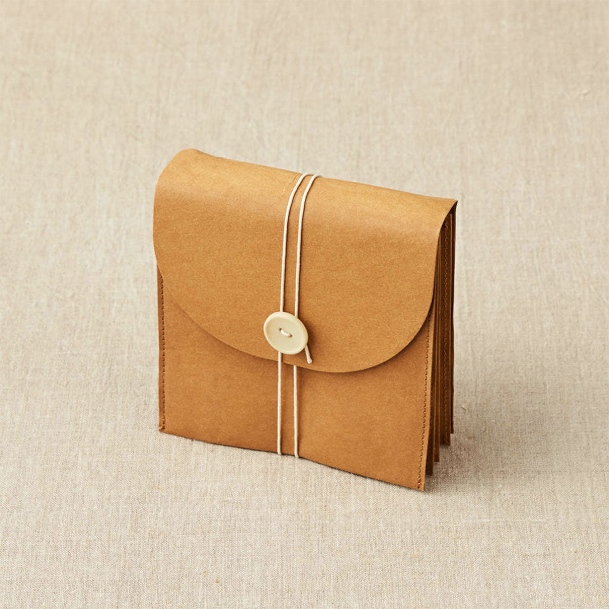 Cocoknits kraft coloured paper based wallet closed with twine and button standing on linen background