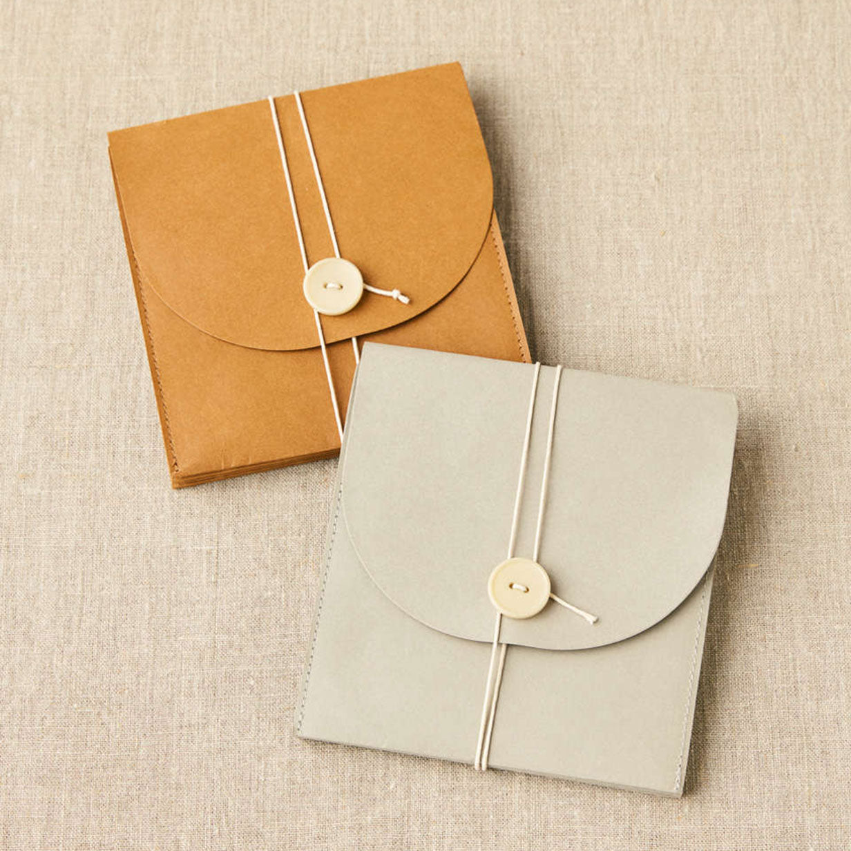 Cocknits paper based project wallets shown lying flat with twine and button closure, kraft and grey colour, placed on linen background
