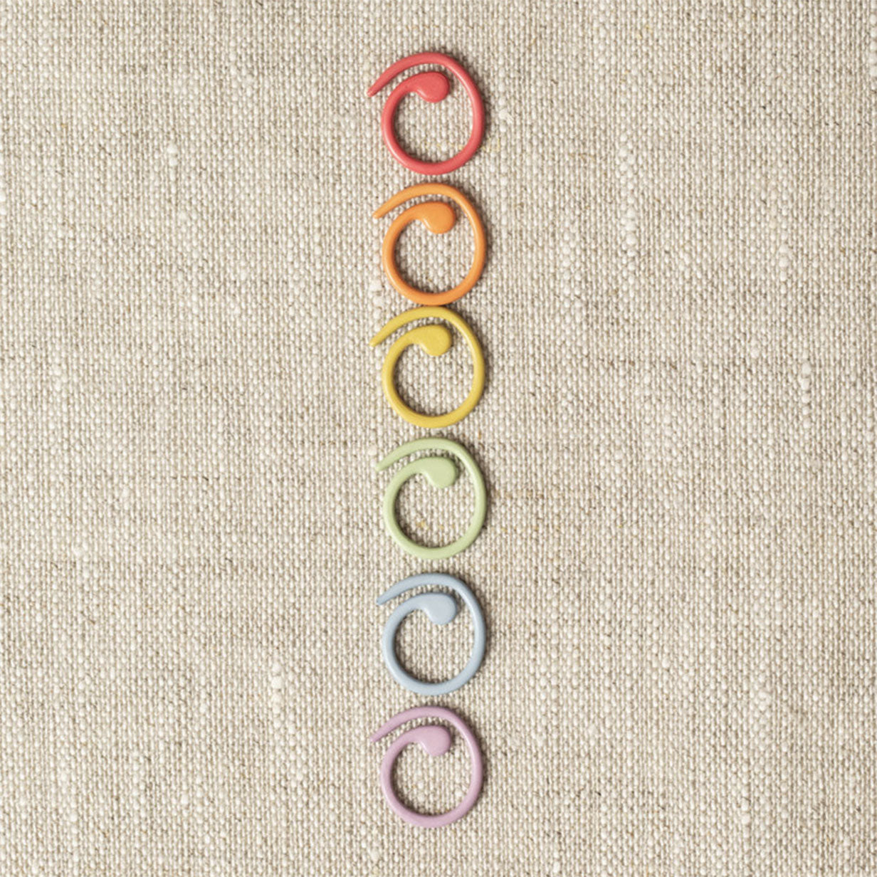 Red, orange, yellow, green, blue and lilac coloured split ring stitch markers placed in a row on linen background