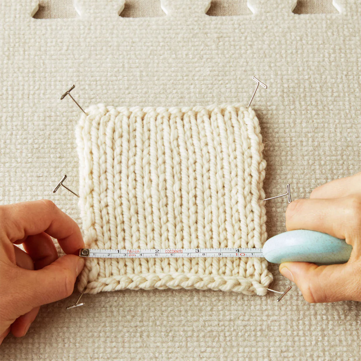 T-pins shown pinned through a swatch of knitting with hands holding a tape measure