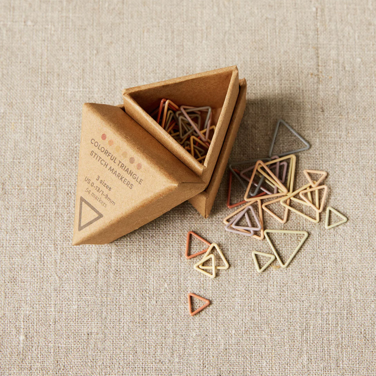 Cocoknits colourful triangle stitch markers in a cardboard box on a linen fabric background