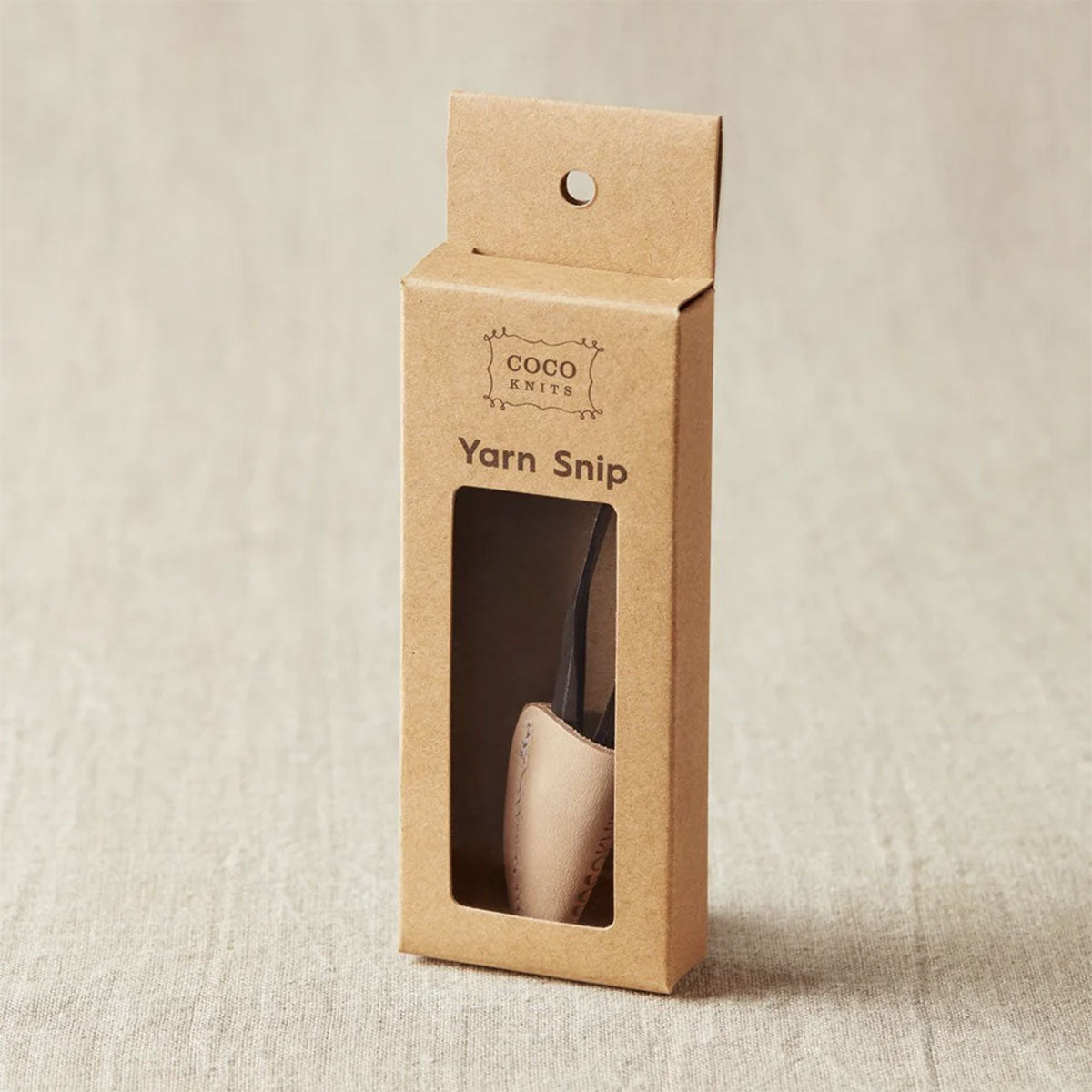 Cocoknits yarn snip in a brown cardboard packaging on a linen fabric background