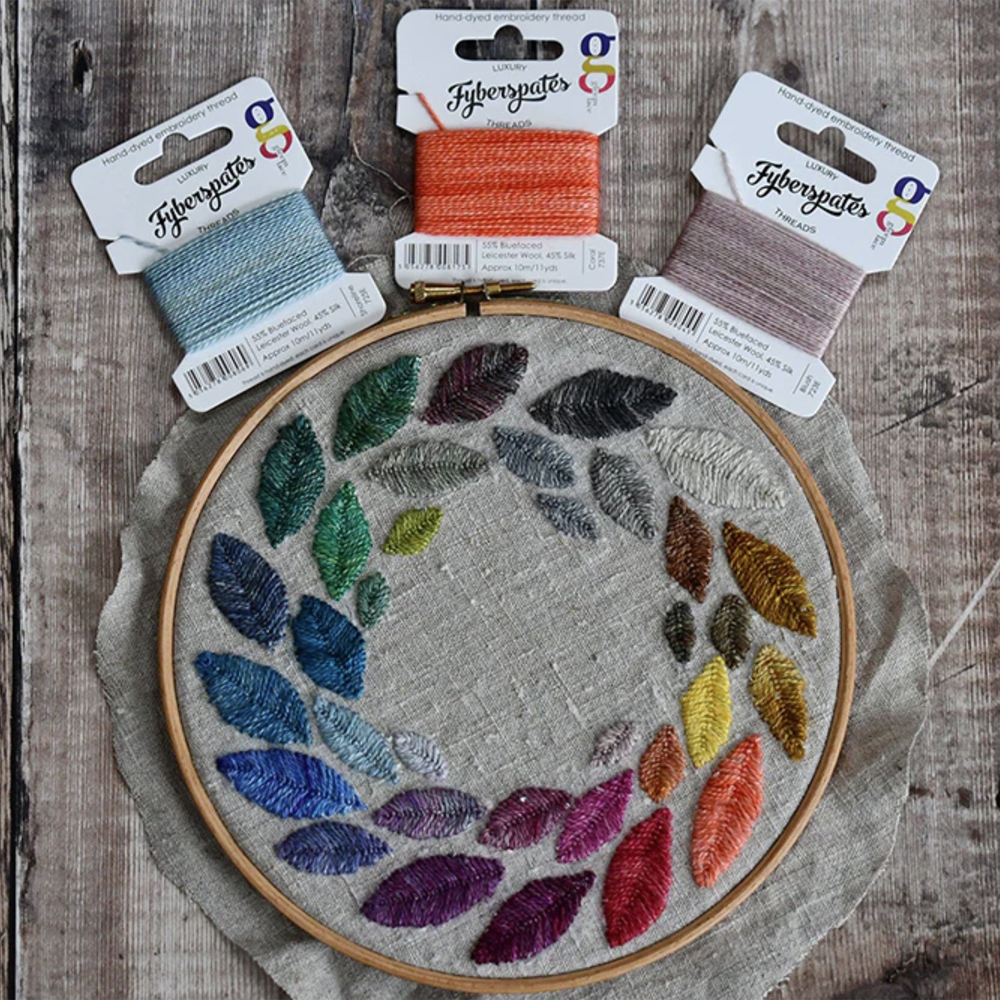 Embroidery hoop with colorful leaf pattern and Fyberspates Gleem Embroidery Thread cards on a wooden surface