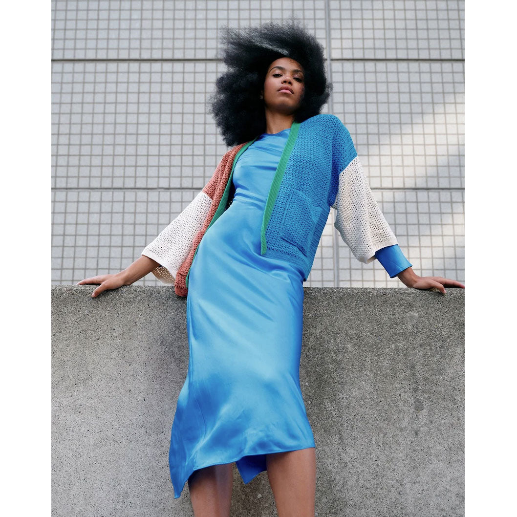 Woman in a blue dress and block colour crochet cardigan leaning against a wall.