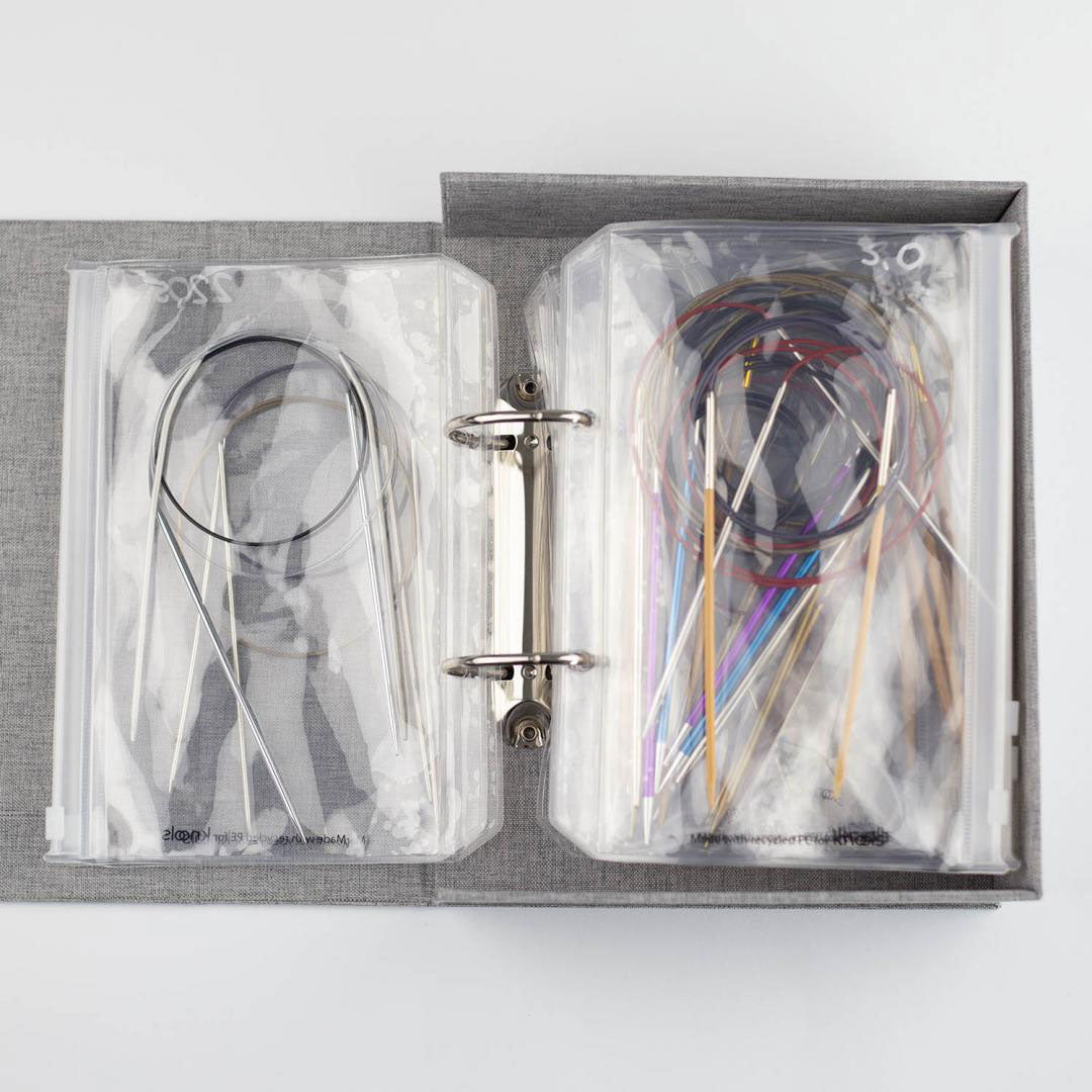 Flat lay view of Knools grey needle garage ring binder folder with clear plastic pockets holding various styles of fixed circular knitting needles 