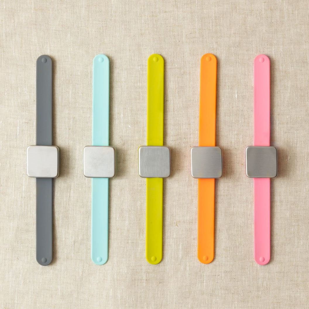 Five colourful wristbands with square magnets in watchface position laid flat on a beige fabric background