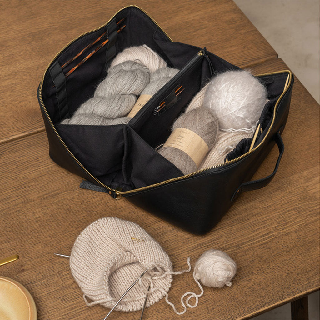 Black leather knitting bag with gold coloured zip, yarn and a knitting project on needles on a wooden table