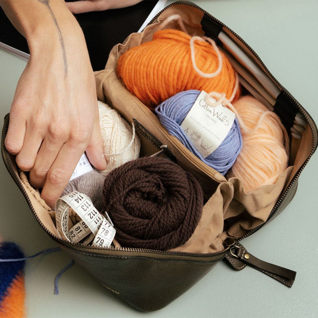 Hand reaching into interior of dark green leather knitting project bag, filled with colourful balls of yarn, crochet hooks and a tape measure placed on a pale green surface