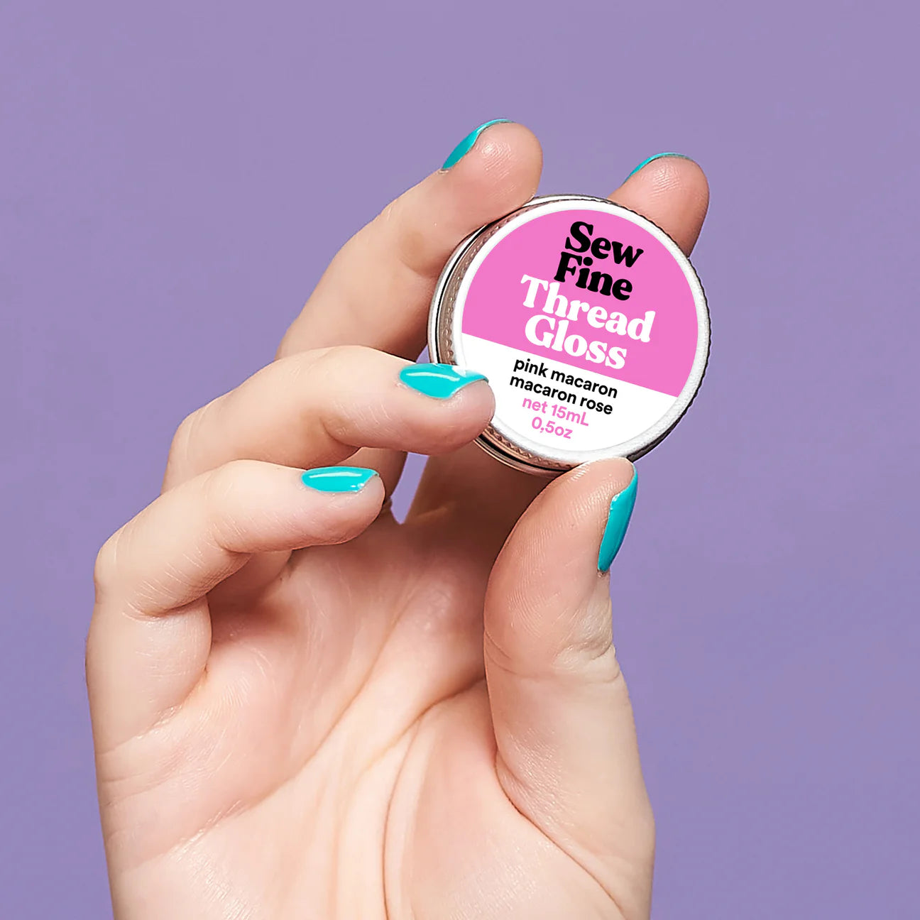 Hand holding a labelled tin of Sew Fine Thread Gloss - Pink Macaron against a purple background