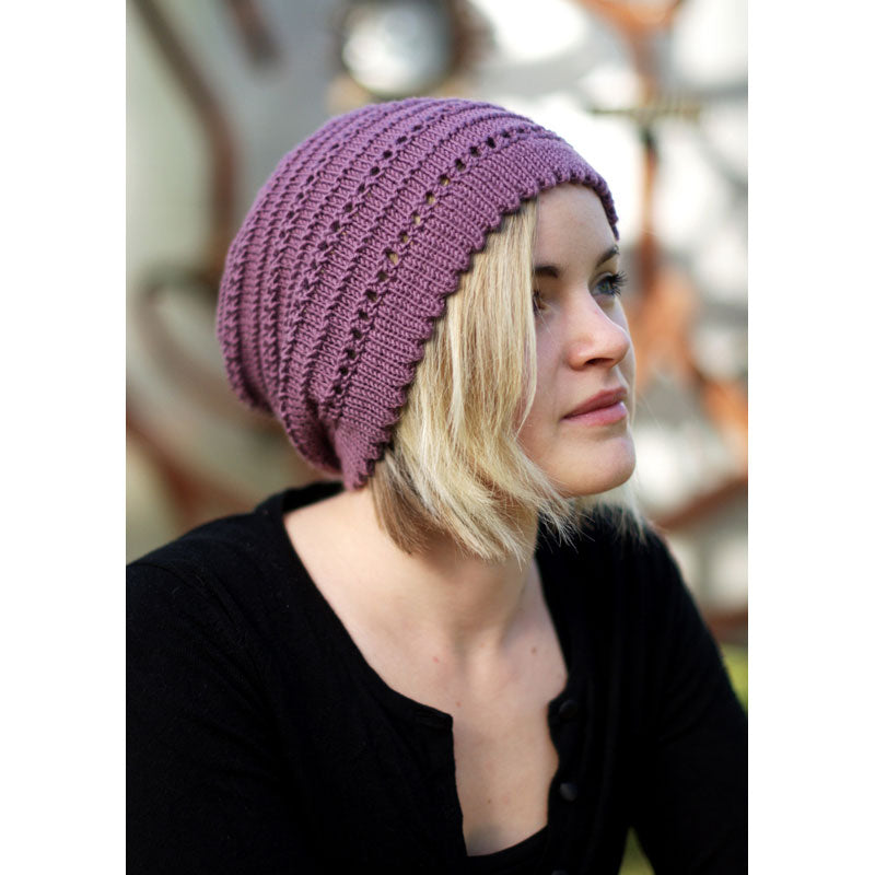 Person wearing purple coloured knitted slouch hat with picot rim, garter stitch contrast and eyelets against a blurred background
