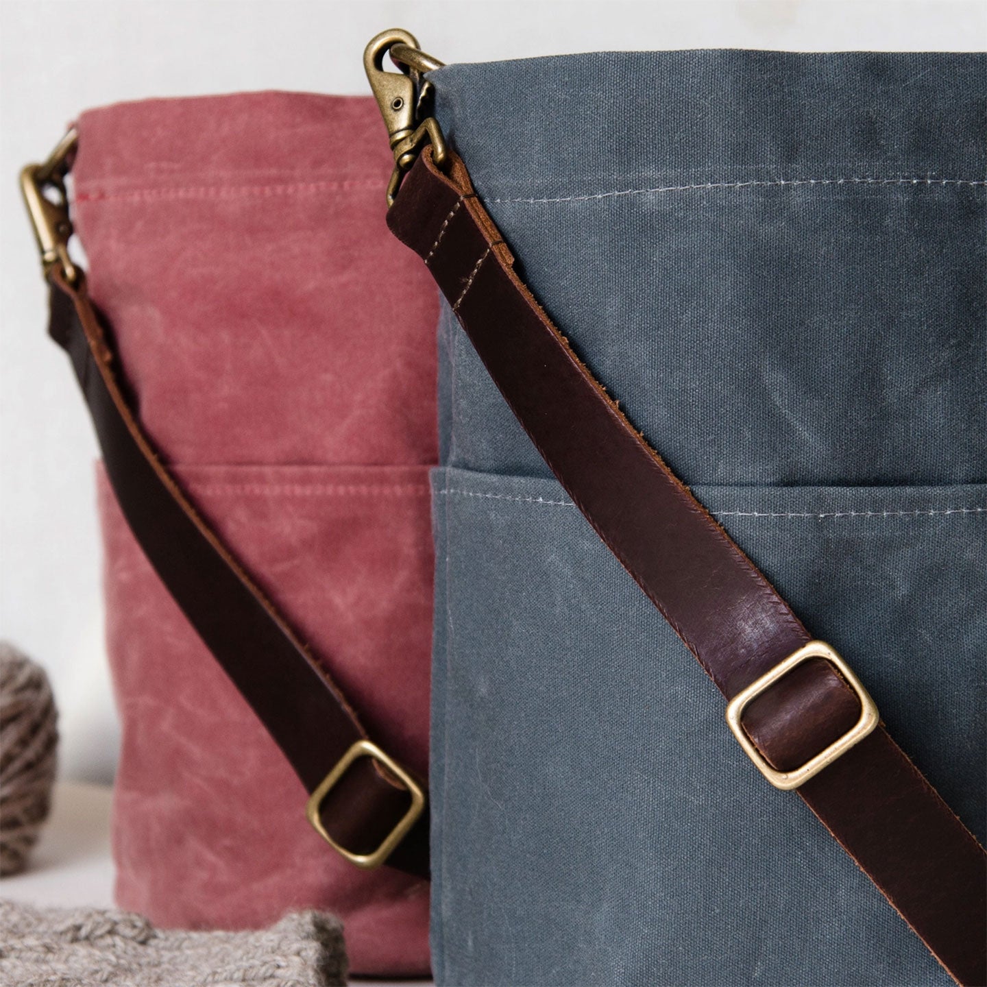 Two waxed canvas bucket bags, one cranberry red and one slate blue, with leather straps and brass buckles on a light background.