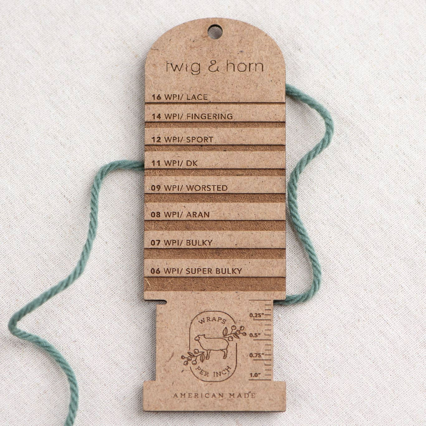 Twig & Horn MDF wraps per inch tool lying across strand of dark green yarn on plain linen background